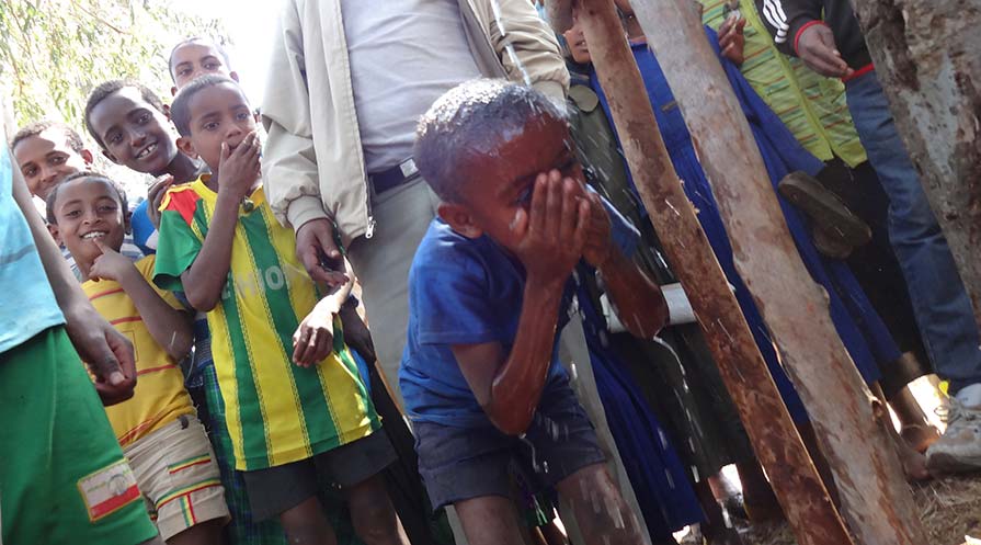 Photo of a child washing his face at an outdoor water pump as other children and adults look on.