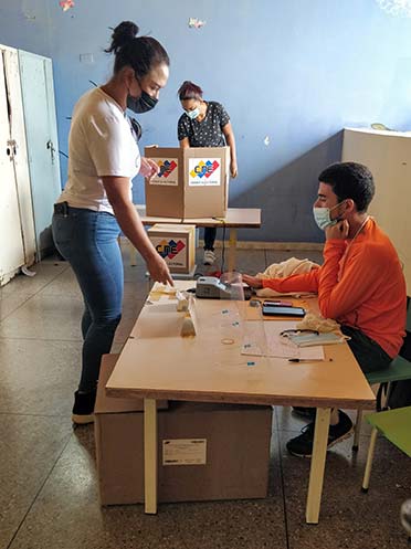 Woman standing in front of a seated poll worker at a polling station in Venezuela.